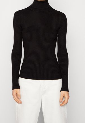 Personne portant un pull à col roulé côtelé noir ajusté et un pantalon blanc ample sur un fond uni.