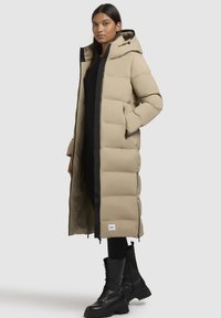 Beige Steppjacke mit Kapuze, Frontreißverschluss und schwarzem Besatz. Verfügt über große Taschen, gestepptes Muster und ist mit schwarzen Kampfboots kombiniert.