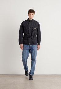 Casaco preto, de tecido pesado, com gola alta, bolsos frontais e um cinto; combinado com jeans de denim azul claro e sapatos pretos.