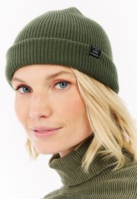 Olijfgroene geribbelde gebreide beanie met een omgevouwen rand en een klein zwart label met "PROTEST HOUSE OF SNOW" dat aan de zijkant is genaaid.