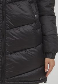 Svart pufferjakke med kviltet tekstur, har full glidelås, ribbestrikkede mansjetter og trykknappdetaljer. Glatt, blank finish.