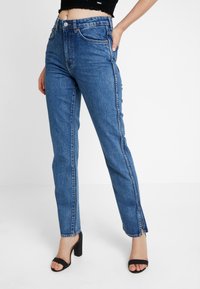 Jean bleu taille haute en denim. Coupe droite avec fentes latérales à l'ourlet. Porté avec des sandales noires à talons.