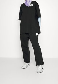 Schwarzes oversized T-Shirt mit V-Ausschnitt und gesticktem Logo, kombiniert mit schwarzen Hosen mit Logoschriftzug, abgerundet mit grauen und weißen Sneakern.