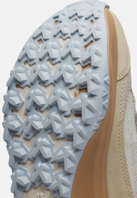 Gros plan sur la semelle beige d'une chaussure avec un motif de traction texturé en forme de X de couleur bleu clair et un détail de semelle intermédiaire marron.