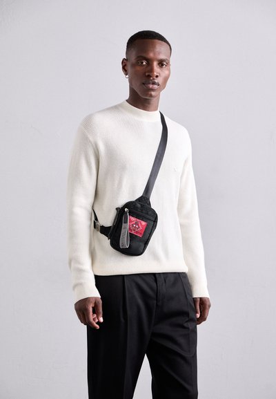 Schwarze Stoffschultertasche mit rotem Patch und grauem Reißverschluss, getragen von einer Person in einem weißen gerippten Pullover und schwarzen Hosen.