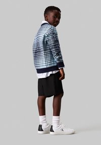 Maglione lavorato a maglia con motivo a quadri blu e blu scuro, camicia bianca sotto, pantaloni corti neri, sneaker bianche con dettagli neri, calzini bianchi al polpaccio.