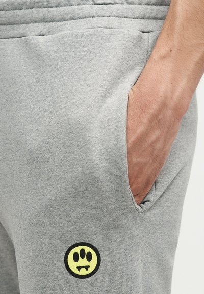 Χέρι στην τσέπη γκρι ανοιχτού χρώματος φόρμας με μικρό κίτρινο και μαύρο λογότυπο πατούσας με κυνόδοντες κοντά στον μηρό.