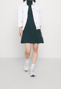 Robe en maille verte avec ourlet évasé, associée à une veste en jean blanche. Chaussures de sport blanches avec des accents gris. Texture lisse et design décontracté.