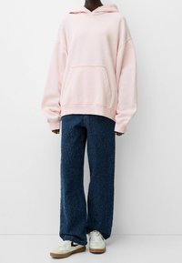 Světle růžová oversized mikina s klokaní kapsou, v kombinaci s modrými širokými kalhotami s texturou a bílými teniskami s černými akcenty.