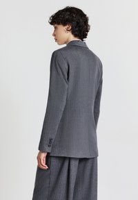 Giovane con capelli ricci corti, che indossa un abito grigio a righe, in piedi di profilo su uno sfondo semplice.