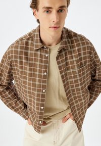 Camisa de cuadros marrones con líneas blancas y rosas claras, llevada sobre un suéter de cuello redondo beige, combinada con pantalones color blanco sucio.