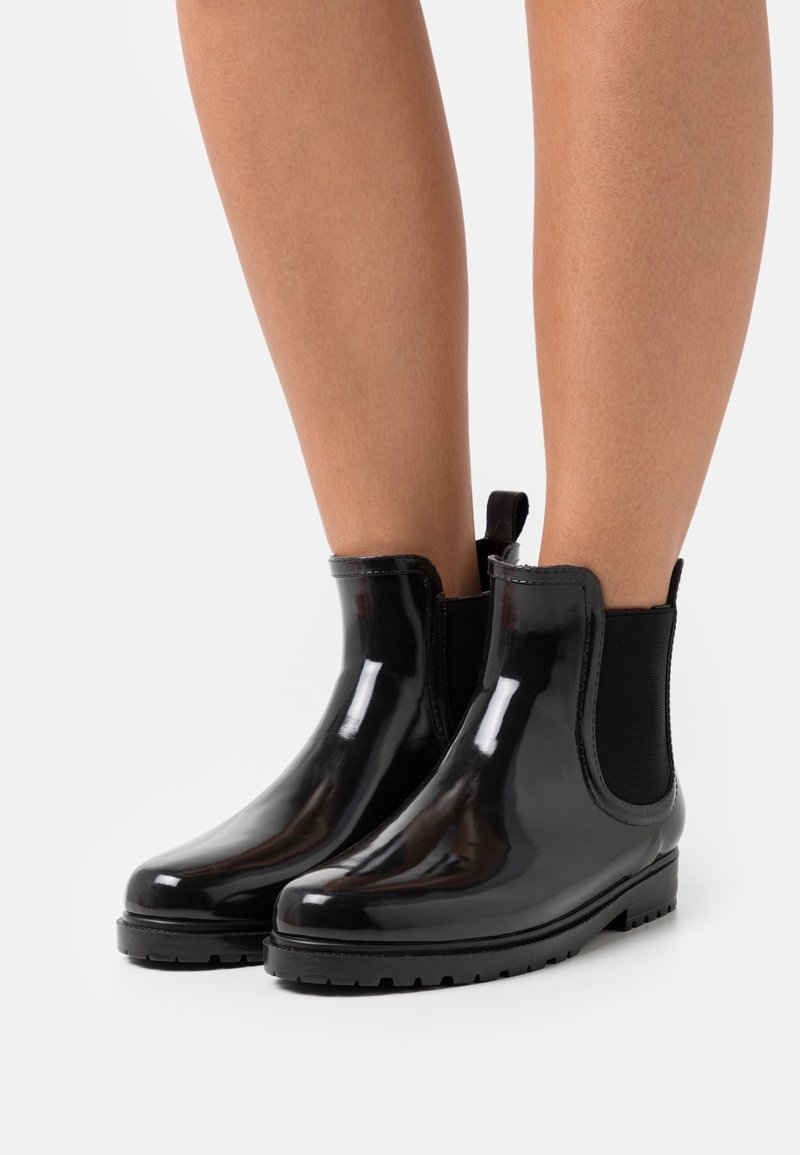 Bottes noires brillantes à la cheville avec panneaux latéraux élastiques portées sur des jambes nues sur un fond clair uni.