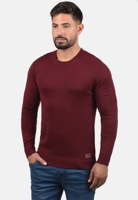 Burgunderfarbener Rundhalspullover im Rippenstrickdesign, mit langen Ärmeln und einem geprägten Lederpatch am Saum. Getragen mit blauen Jeans.