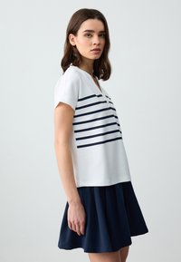 Weißes T-Shirt mit kurzen Ärmeln und navyfarbenen horizontalen Streifen, mit V-Ausschnitt und lässiger Passform, kombiniert mit einem navyfarbenen ausgestellten Rock. Glatte Textur.