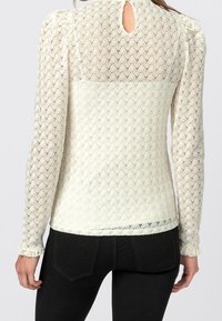 Långärmad blus i krämfärg med ett strukturerat spetsmönster. Har en nyckelhålsglenn och volangärmar. Fitted design med en mjuk och lätt känsla.