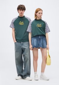 Νεαρός άντρας και γυναίκα στέκονται δίπλα-δίπλα, φορώντας ταιριαστά πράσινα μπλουζάκια με κοντά μανίκια και σχέδια, τζιν παντελόνια, λευκά αθλητικά παπούτσια και κρατώντας μια κίτρινη τσάντα χειρός.