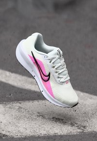 Chaussure de sport légère avec une tige en mesh blanc respirant, des accents roses, un logo swoosh noir et un col rembourré avec une épaisse semelle blanche.