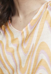 Top in lino a scollo a V con un motivo ondulato giallo e viola chiaro su uno sfondo crema, che mette in risalto una texture morbida e un design fluido.