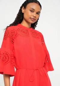 Vestido vermelho com recortes decorados em bordado nas mangas curtas, gola alta e um cinto atado na cintura. Textura de tecido suave.