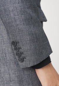 Manche de blazer gris texturé avec quatre boutons noirs, comportant un léger revers dévoilant un détail en maille sombre en dessous.
