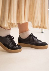 Musta nahka- ja mokkanahkakengät, joissa on nyöritys ja korotettu pohja, yhdistettynä vaaleisiin sukkiin. Hameessa on laskostettu muoto ja raidallinen kuvio.