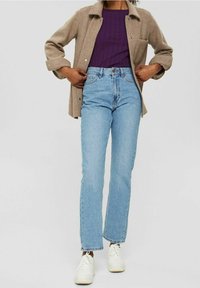 Höga ljusblå jeans som matchas med en lila ribbstickad topp och en beige knäppt skjorta med ficka och avslappnad passform. Vita sneakers.