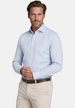 Mann mit hellbraunen Haaren, der ein hellblaues Hemd mit Knöpfen, beige Hosen und einen dunkelbraunen Gürtel trägt, steht vor einem schlichten Hintergrund.