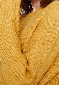 Žlutý pletený svetr s hrubou texturou. Má volný střih a žebrované detaily na manžetách a lemu. Měkký, teplý materiál.