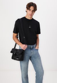 Črna majica s kratkimi rokavi s kvadratno logo nalepko, v kombinaciji z svetlo modrimi jeans hlačami in črno platneno torbo za čez ramo z zapiralom.