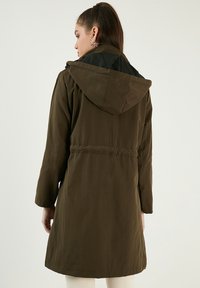 Jachetă cu glugă de culoare verde măsliniu, cu talie strânsă, mâneci lungi și un material neted, ușor; are un căptușeală neagră.