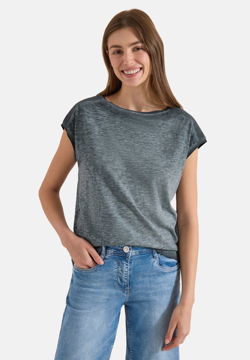Jeune femme aux longs cheveux bruns, souriante, portant un haut gris à manches courtes et un jean bleu clair, une main dans la poche.