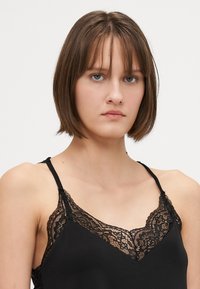 Svart camiset med blondekant og tynne stropper, laget av mykt stoff med intrikate blonde detaljer ved halsen.