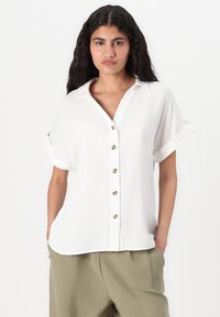Camicia bianca a maniche corte con risvolti e bottoni dorati; tessuto morbido con vestibilità rilassata, abbinata a pantaloni larghi di un verde chiaro.