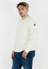 Maglione bianco a coste con colletto rotondo, bordo beige a contrasto e toppa con il logo sulla manica sinistra; indossato con jeans blu.