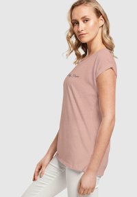 Lys rosa t-skjorte med kort arm laget av mykt stoff, med rund hals og brodert tekst på forsiden, kombinert med hvite bukser.