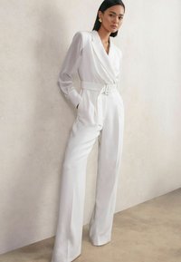 Balts pielāgots kombinezons ar gariem piedurknēm, platajām biksēm, jostas detaļu un gludu struktūru, saskaņots ar lielām apaļām auskarām.