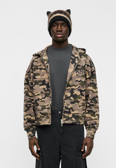 Sweat à capuche zippé camouflage en beige, olive et noir avec poches avant ; associé à un t-shirt gris foncé et un bonnet en maille noir avec des oreilles.