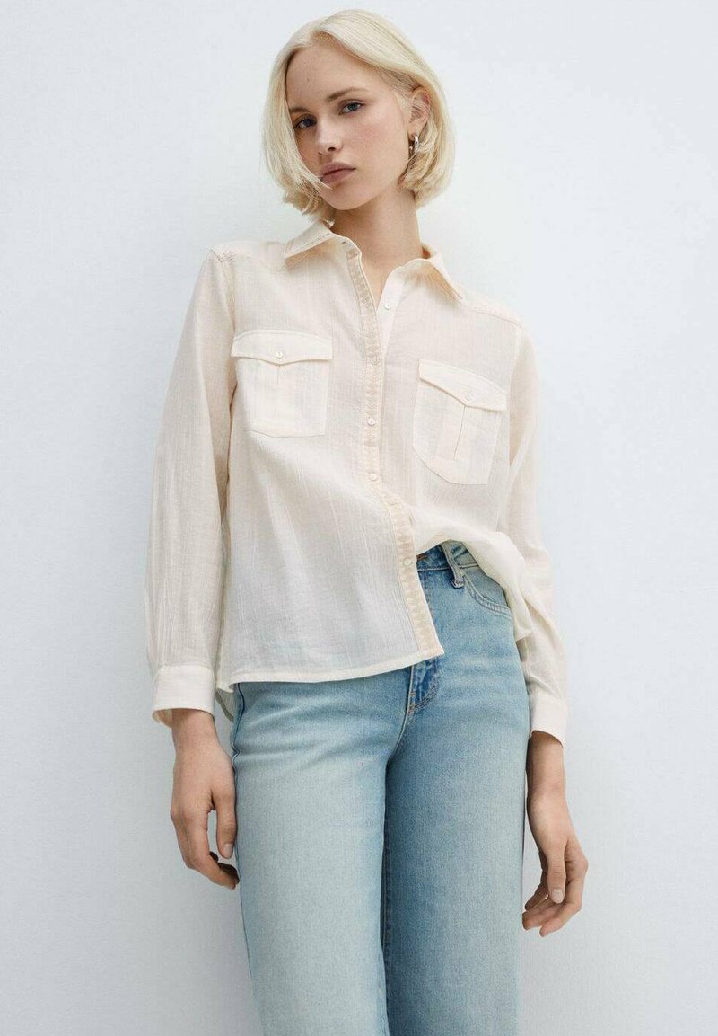 Camicia button-up beige chiaro realizzata in un tessuto morbido, con due tasche sul petto, maniche lunghe e una vestibilità comoda, abbinata a jeans azzurri chiari.