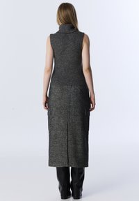 Vrouw die met de rug naar ons staat, gekleed in een mouwloze donkergrijze coltrui, een lange grijze rok met zakken en zwarte laarzen tegen een effen achtergrond.