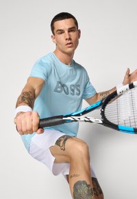 T-shirt de sport bleu clair avec un logo "BOSS" noir audacieux, associé à un short blanc ; la personne tient une raquette, mettant en valeur de prominents tatouages.