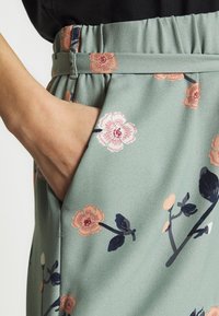 Světlé zelené květinové kalhoty s elastickým pasem. Mají růžové a námořnické modré květinové vzory, boční kapsy a hladký textil.