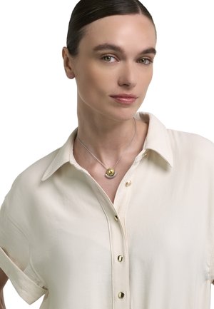 Žena s zalizanom tamnom kosom, odjevena u kremastu kratkih rukava košulju s kopčanjem i srebrnu ogrlicu sa zlatnim kuglastim privjeskom.