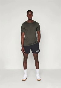 Olíva zöld, rövid ujjú ing és tengerészkék sort, fehér "BORG" logóval, fehér sportcipővel viselve, sima háttér előtt állva.