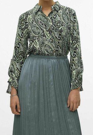 Femme portant un chemisier vert à motif paisley rentré dans une jupe plissée taille haute vert atténué.