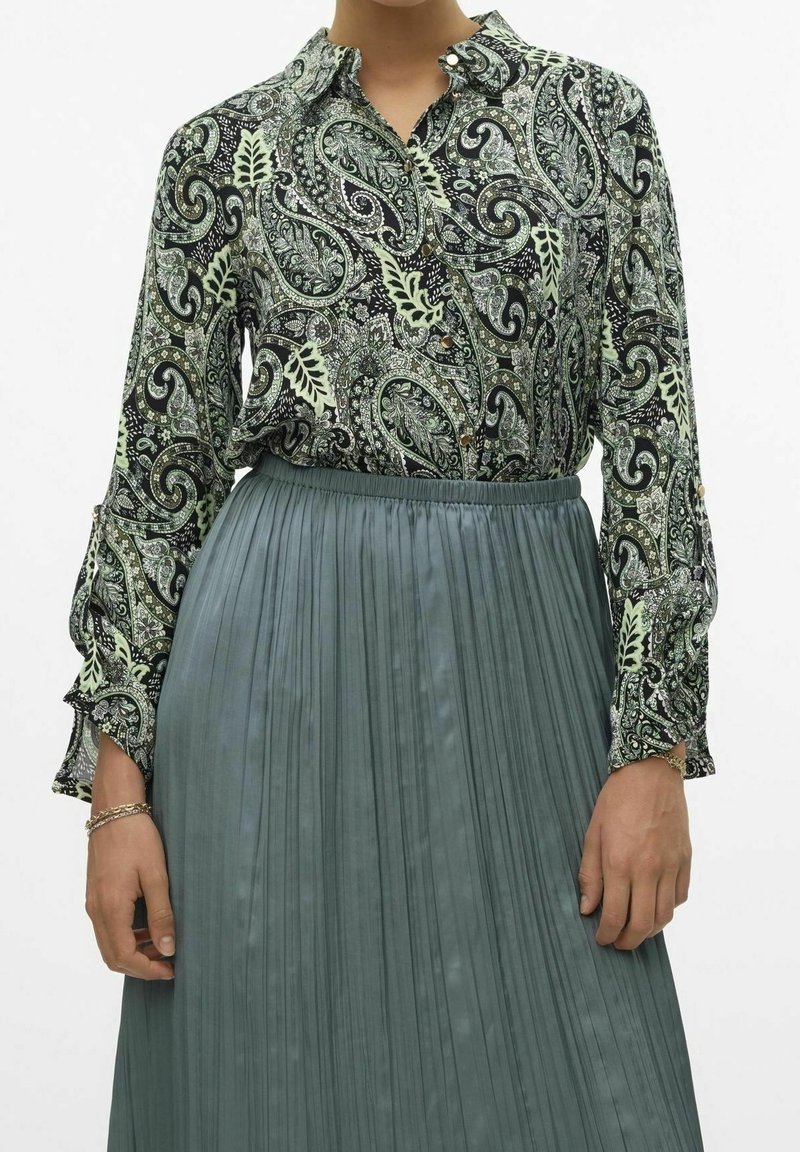 Femme portant un chemisier vert à motif paisley rentré dans une jupe plissée taille haute vert atténué.
