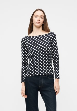 Kvinde iført en sort bluse med lange ærmer og hvide polkaprikker samt mørkeblå jeans, stående foran en ensartet lys baggrund.