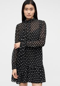 Νεαρή γυναίκα με μακριά σκουρόχρωμα μαλλιά, φοράει μαύρο διάφανο φόρεμα με πουά και δεμένη κορδέλα στο λαιμό, στέκεται μπροστά σε απλό φωτεινό φόντο.