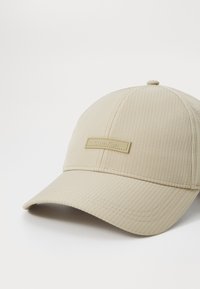 Tan baseball cap with vertical ribbed texture, curved brim, and a small rectangular logo label on the front.