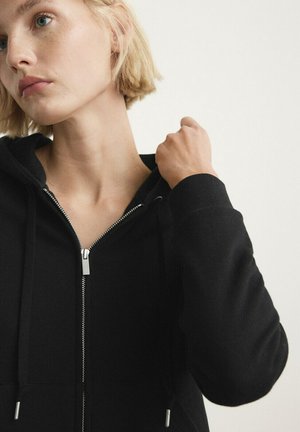 Persona joven con cabello corto y rubio, que lleva una sudadera negra con cremallera, sosteniendo el cordón de la capucha cerca del hombro sobre un fondo liso.