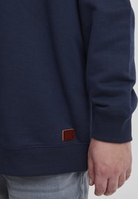 Felpa blu navy realizzata in morbido cotone, con polsini e orlo a costine, dotata di una toppa in pelle marrone con logo nella parte inferiore dell'orlo.